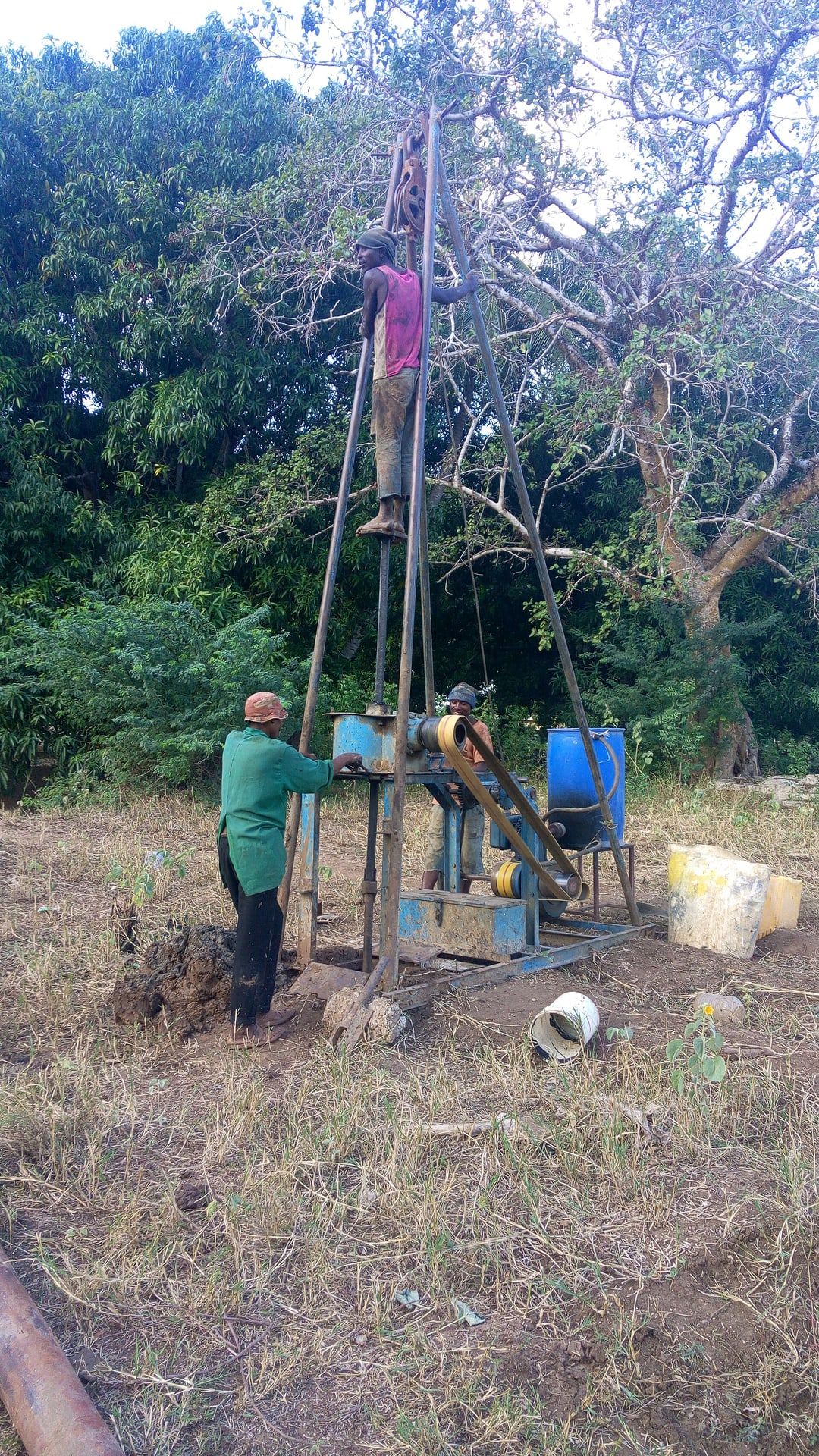 Three men drilling a borehole with a hand-operated tripod rig and a small engine, trees behind.