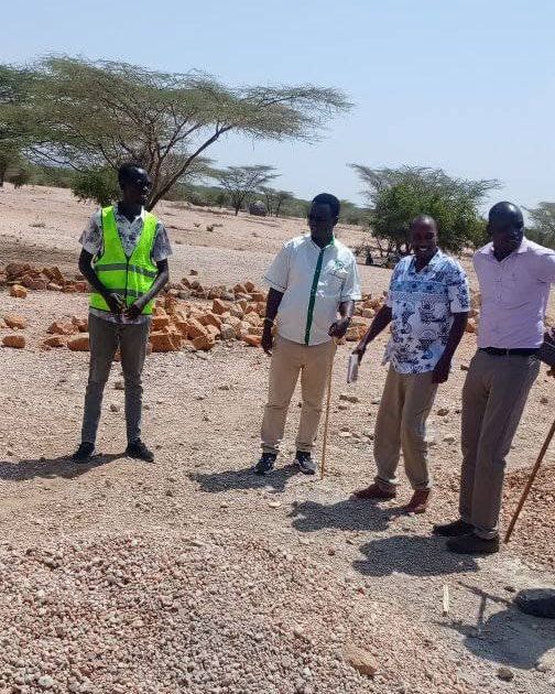 Four people standing on a building site by piles of gravel and stone, acacia trees behind.