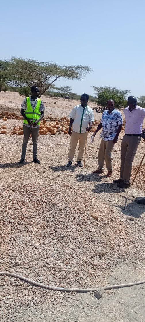 Four people standing on a building site by piles of gravel and stone, acacia trees behind.