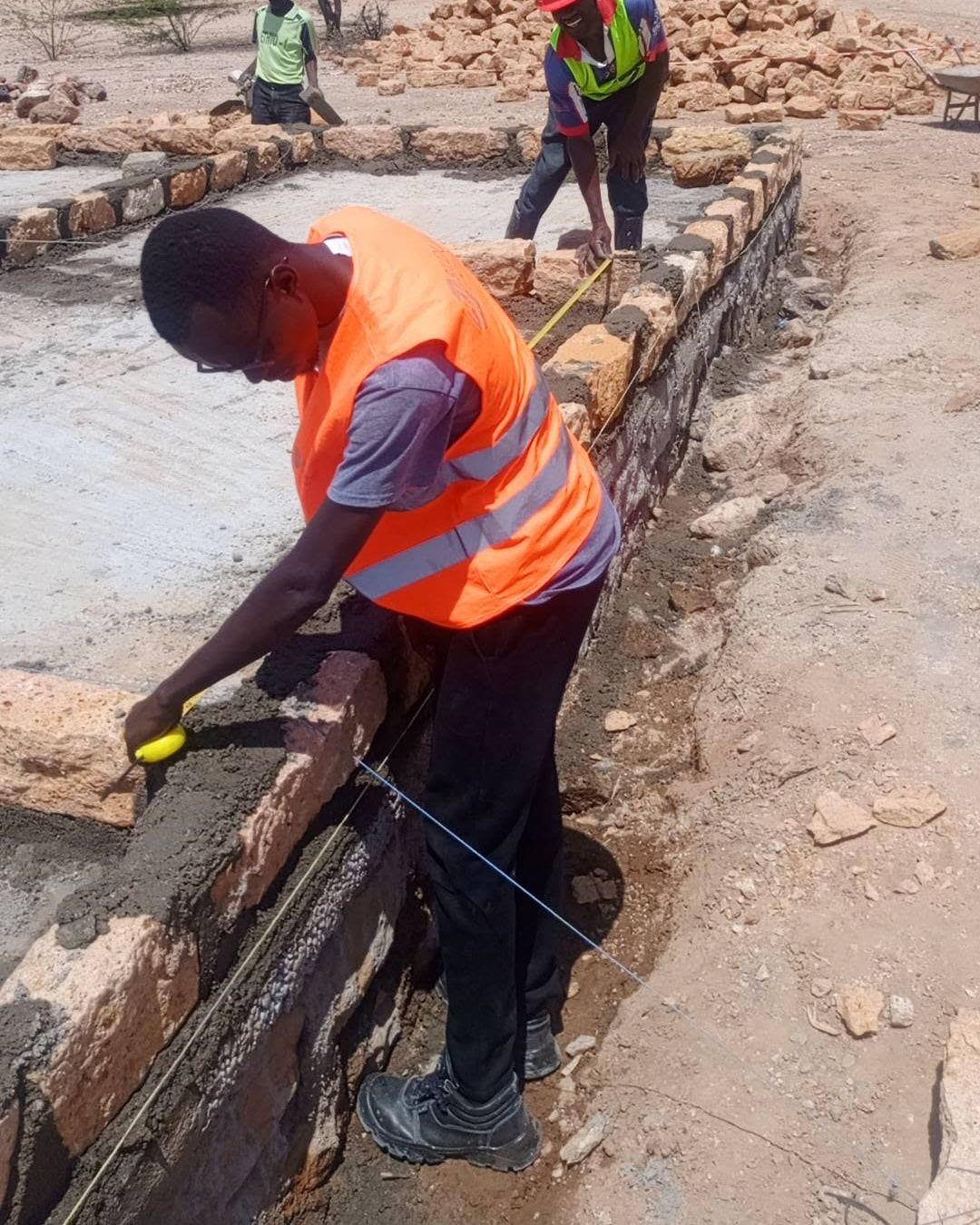An engineer in an orange vest checking a stone foundation course with a tape measure, a colleague in a red hard hat behind.