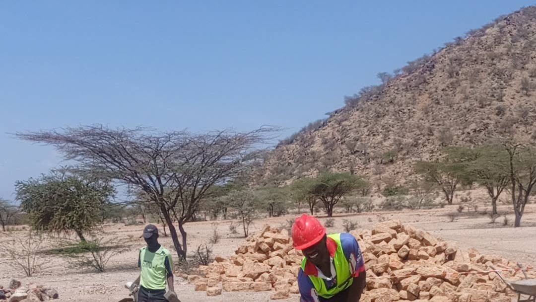 An engineer in an orange vest checking a stone foundation course with a tape measure, a colleague in a red hard hat behind.