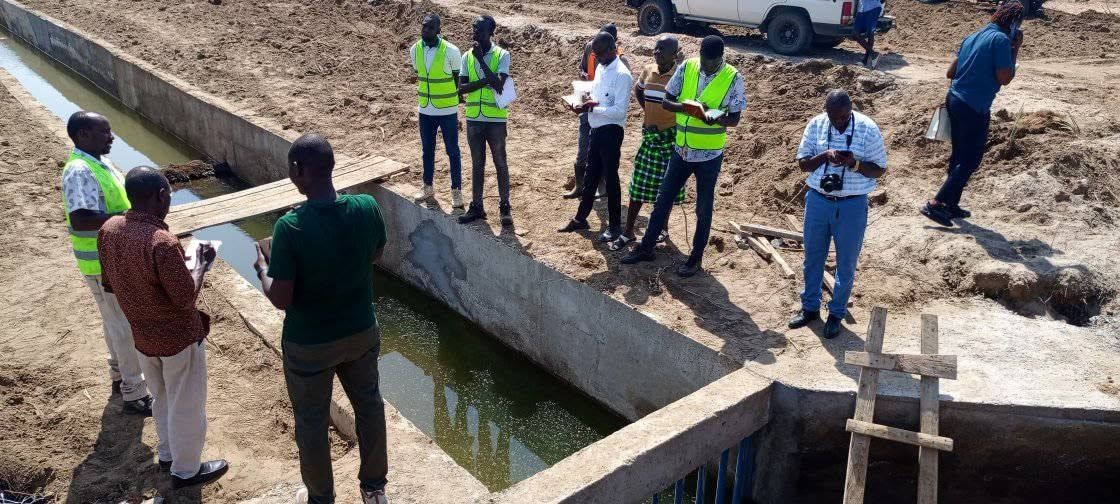 Engineers with clipboards inspecting a concrete-lined canal at Napak, a pickup parked behind.