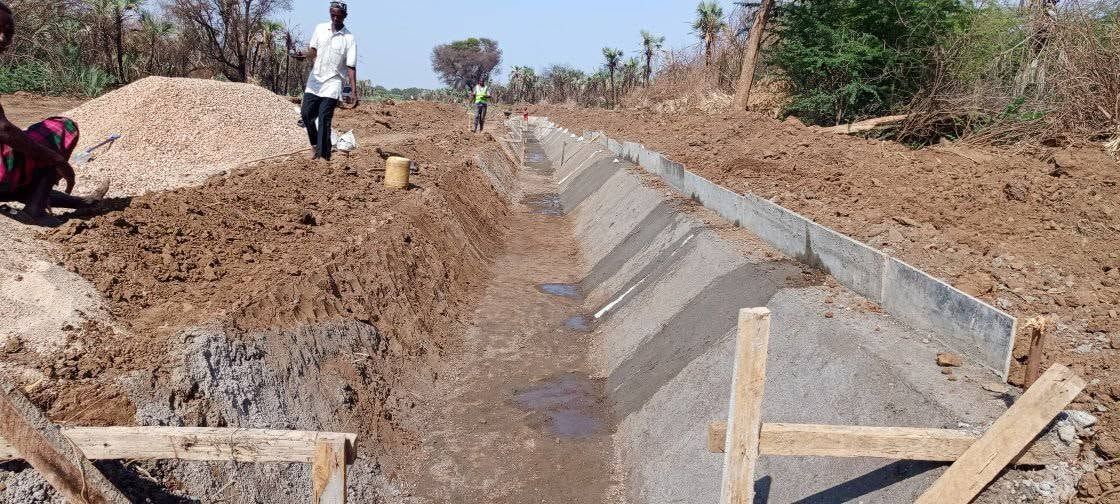 A canal under construction at Napak: one bank lined in concrete, the other still earth, with doum palms behind.